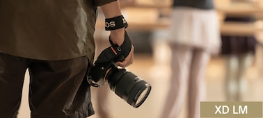 Usage image showing a man carrying in one hand, apparently effortlessly, a camera body with the lens mounted and wrist strap attached